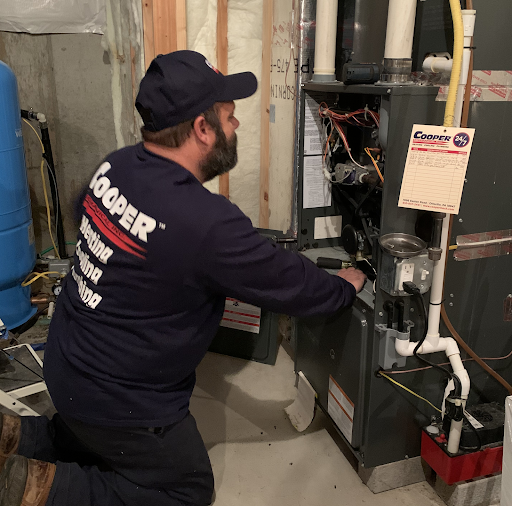 An HVAC technicians performs maintenance on a heating system. He is wearing a Cooper Mechanical shirt and hat.